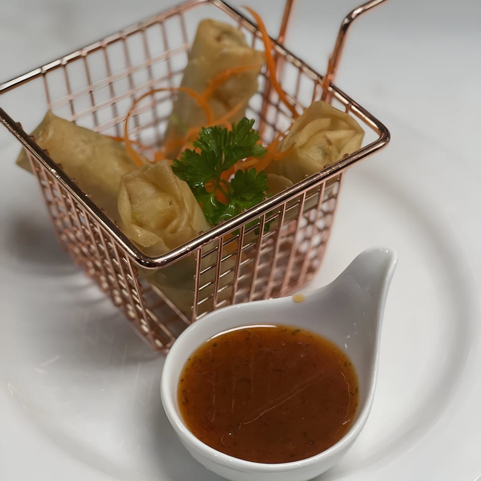 Crispy spring rolls in a rose gold basket with a side of dipping sauce on a white plate.
